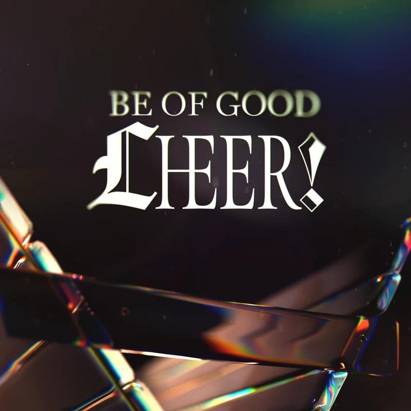 The text 'BE OF GOOD CHEER!' in a stylized white font on a dark background, seen through a crystalline object that refracts rainbow light in the foreground.